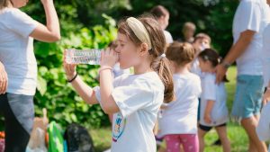 Young girl drinking water during sunny outdoor activity with friends in a park setting.